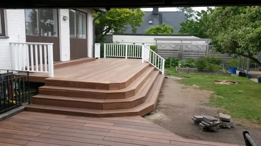 Brunt träträdäck med bred trappa och vitt balustradräcke intill en vit villa, omgivet av grön trädgård.