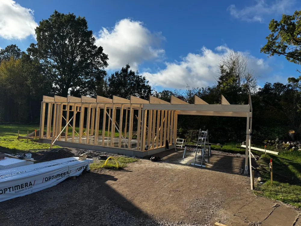 Träregelstomme till en ny byggnad under konstruktion mot blå himmel, med Optimera-material synligt på arbetsplatsen.