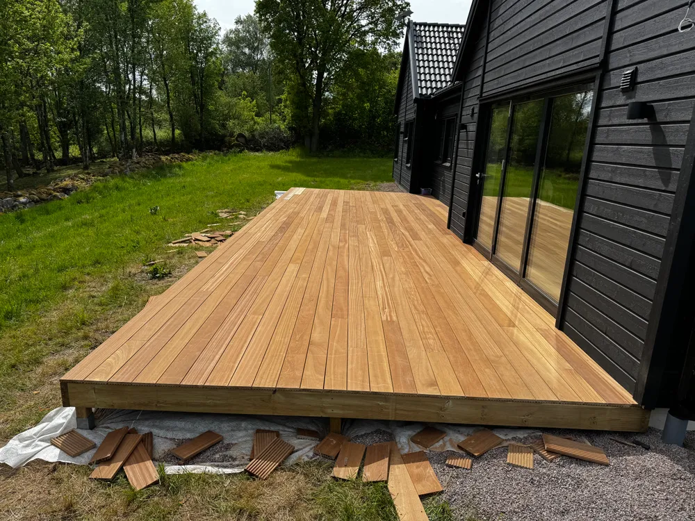 Nybyggt ljust träträdäck intill ett modernt svartmålat hus med stora glasdörrar, omgivet av grön gräsmatta och träd.