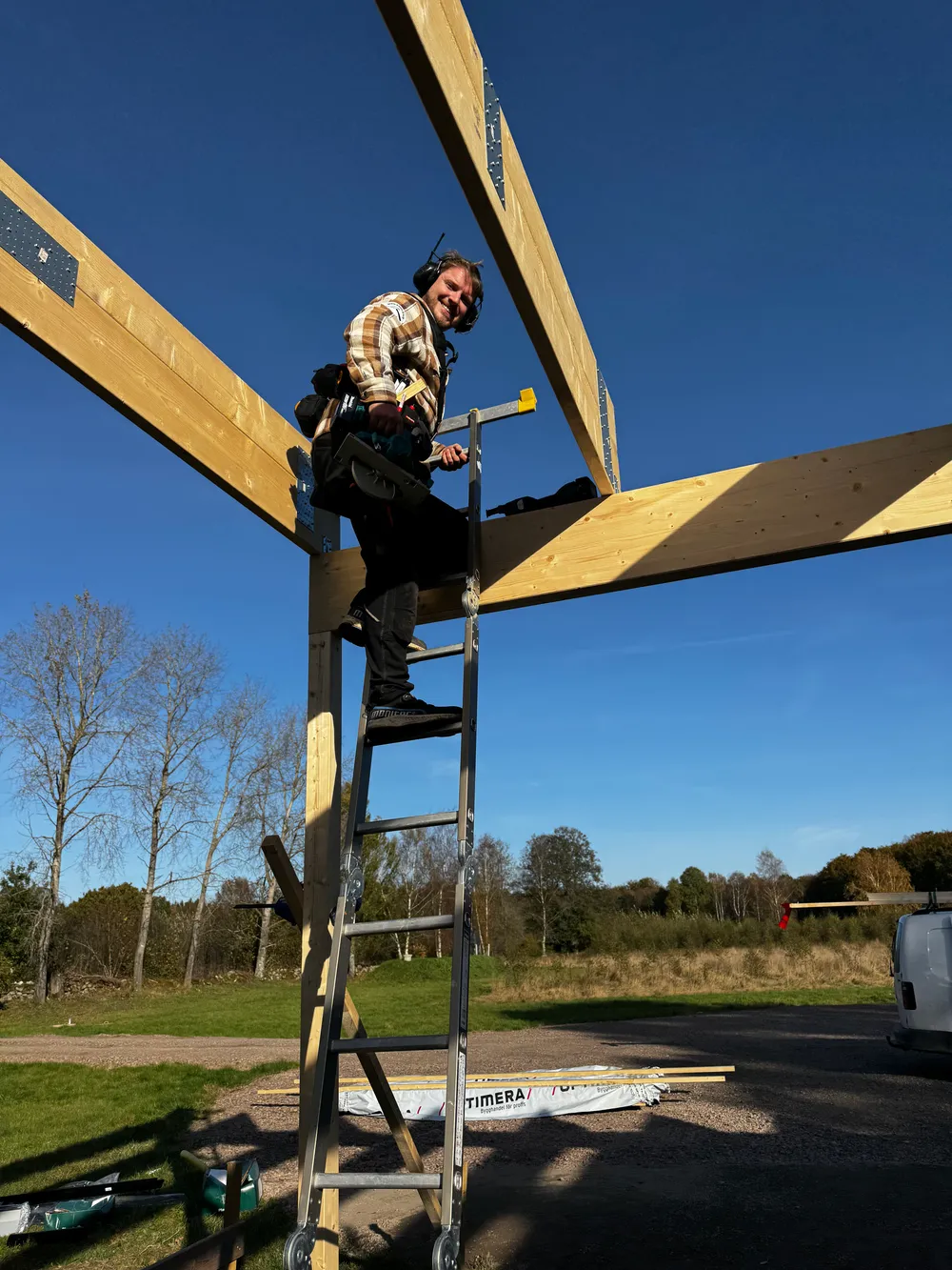 Snickare klättrar upp för en stege mellan stora träbalkar på en byggarbetsplats under en klarblå himmel.