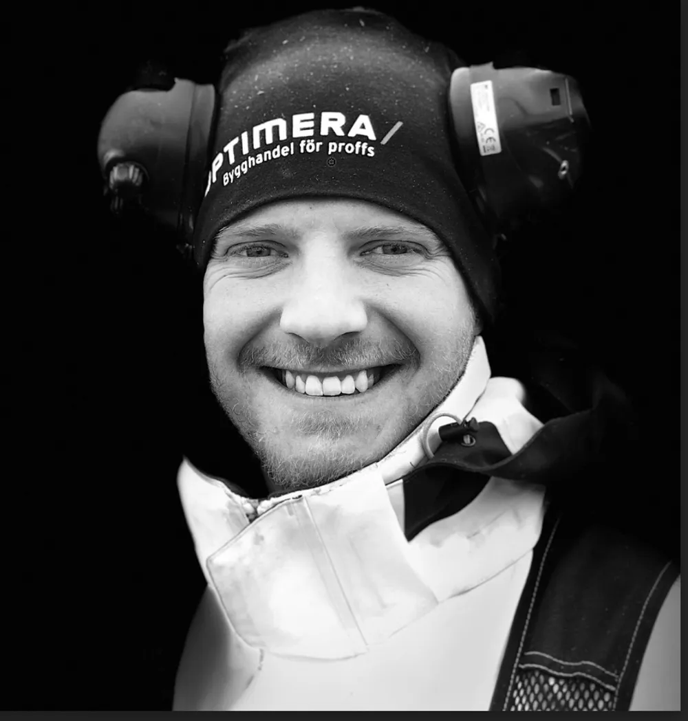 Svartvitt porträttfoto av snickare Filip från Skånska Snickaren, iklädd hörselskydd med ett leende.