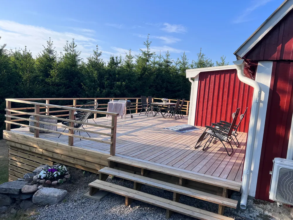 Stort träträdäck med horisontellt räcke och utemöbler intill en röd ladugård under en blå sommarhimmel.