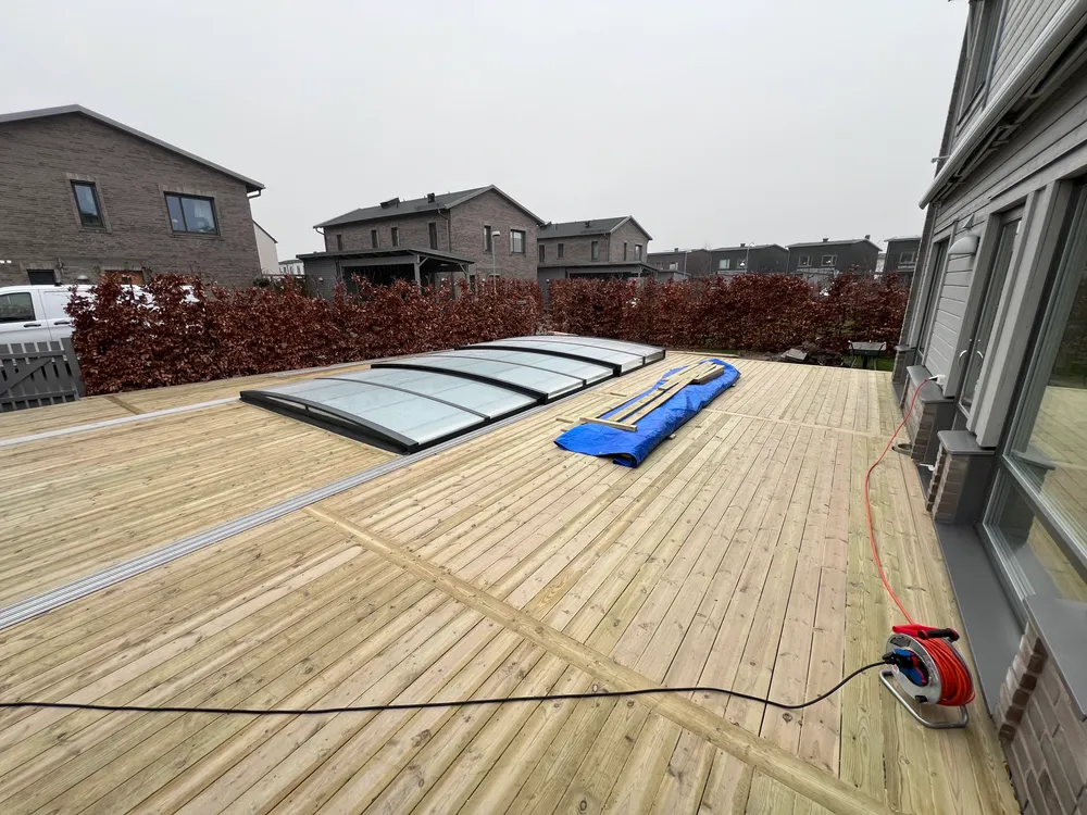 Nybyggt träträdäck runt en poolöverbyggnad i ett villaområde med grå husbebyggelse i bakgrunden.