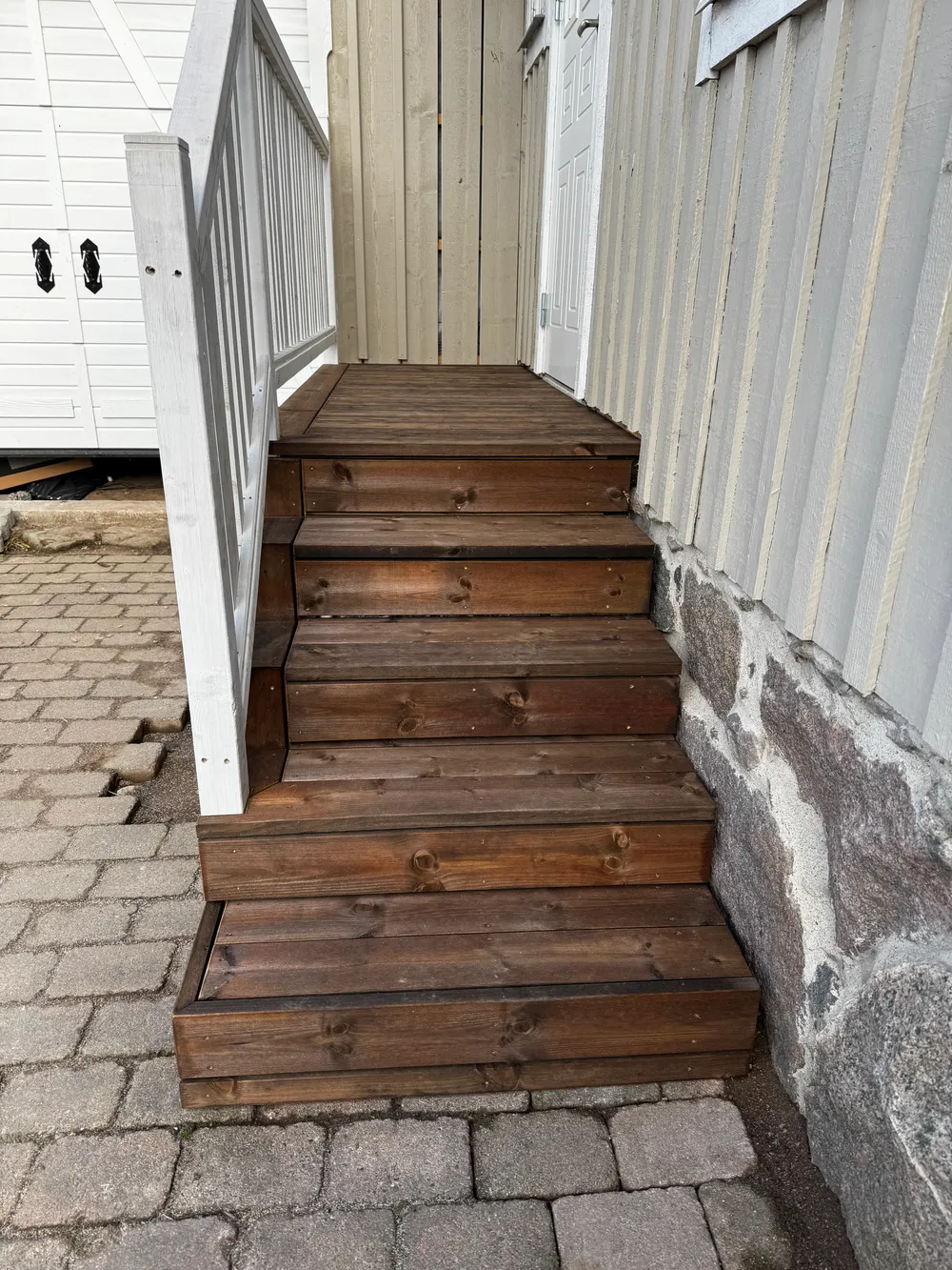 Mörkt brunbetsad vertikal träpanel som fasadbeklädnad på en byggnad, med synlig stenmur och vit garageport.